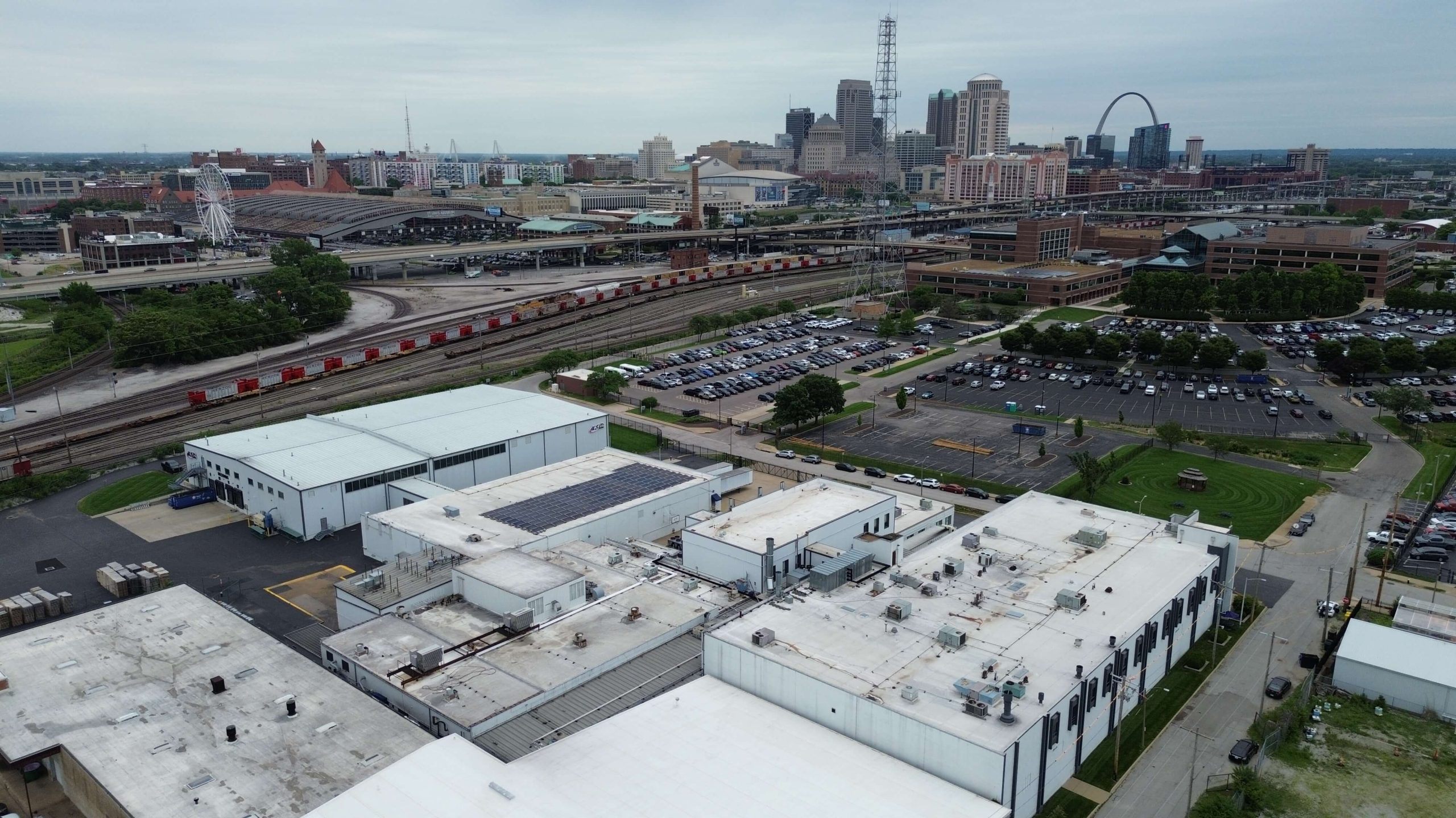 Aerial view of an industrial area with white-roofed buildings, rail tracks, parking lots, and a city skyline with the Gateway Arch in the background.