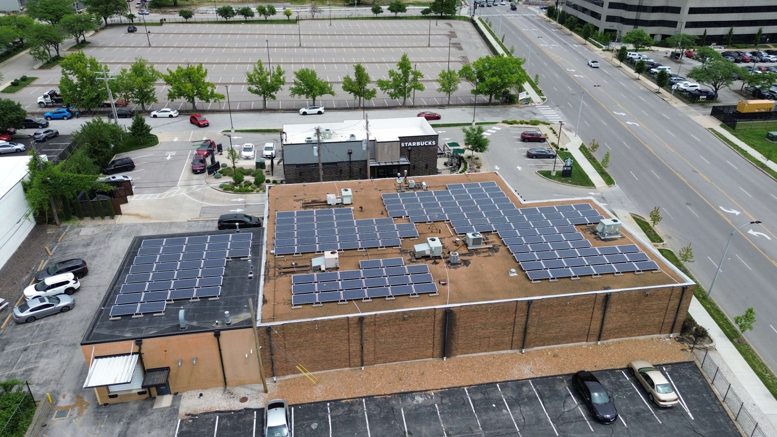 Aerial view of a flat-roofed commercial building with multiple solar panel arrays, surrounded by parking lots, trees, and adjacent roads.