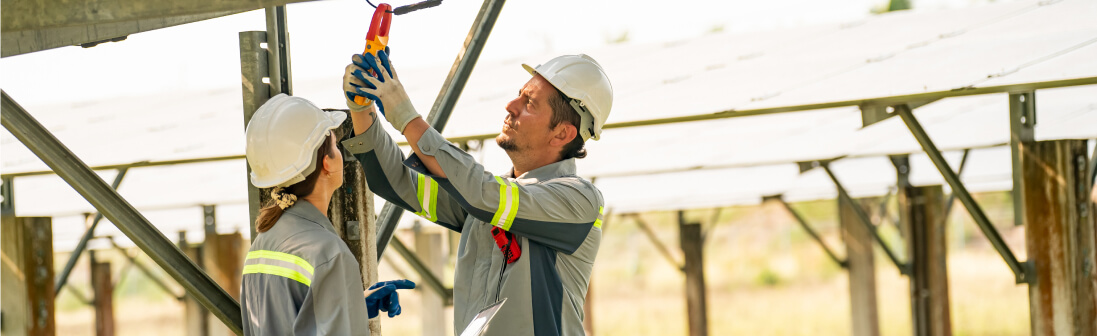 Two commercial solar installers wearing helmets and safety gear install or repair electrical equipment on a solar panel structure outdoors.