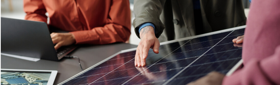 Three people in business attire discuss and examine a solar panel on a desk, representing commercial solar installers, with a laptop and documents visible nearby.