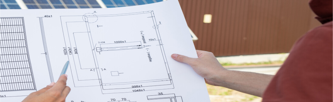 Two people hold and discuss a technical architectural blueprint outdoors, with part of a solar panel and a brown building visible in the background, highlighting the expertise of commercial solar installers at work.