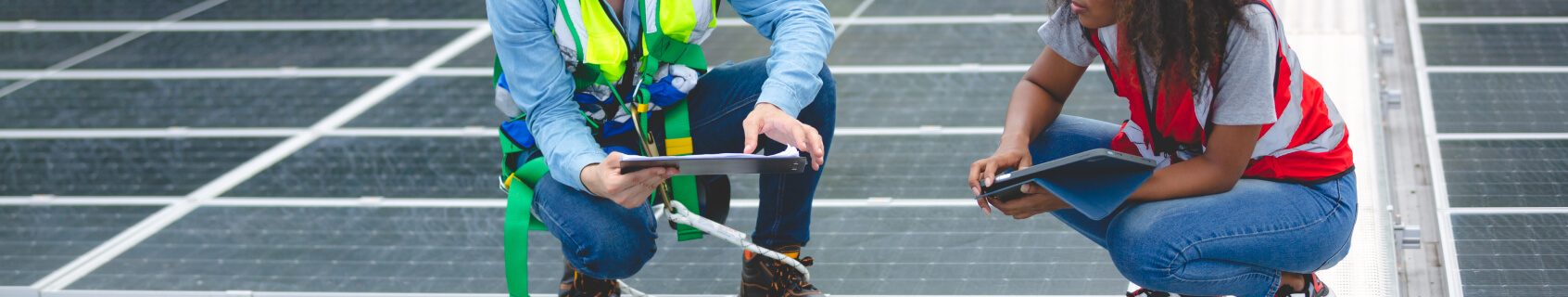 Two workers wearing safety gear crouch on a rooftop during a solar panel installation, holding a clipboard and tablet while inspecting the setup.