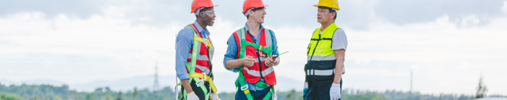 Three construction workers wearing safety vests and helmets stand outdoors, talking and holding equipment on a solar panel installation work site.