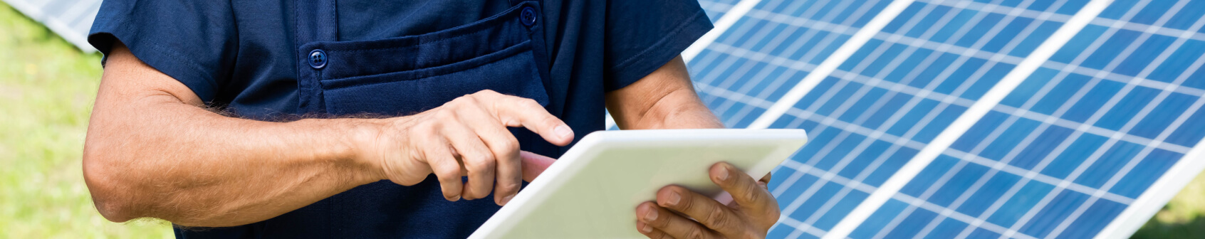 A person in a blue uniform uses a tablet while standing in front of a solar panel installation outdoors.