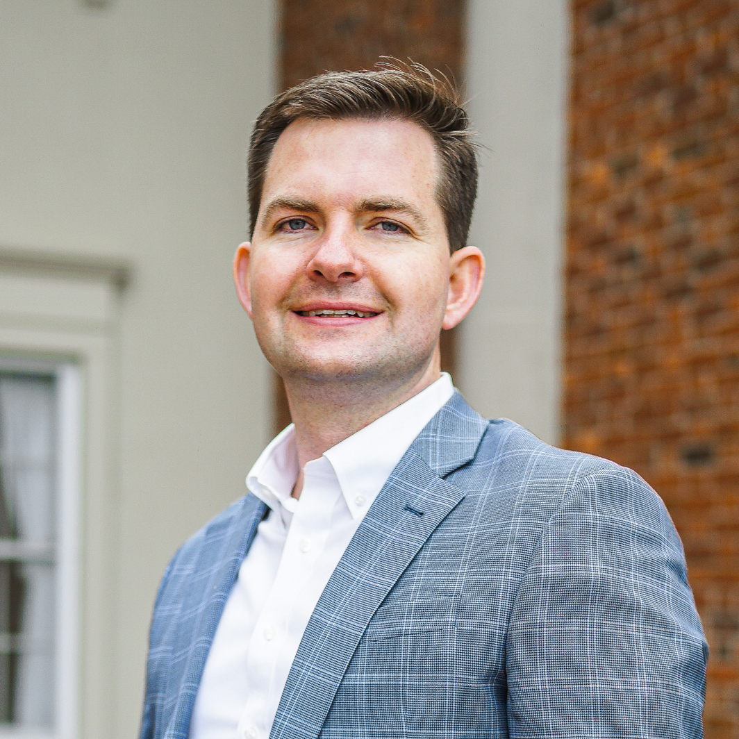 A man in a light blue checked suit jacket and white shirt stands in front of a building with brick and light-colored walls, smiling at the camera, representing Brightergy renewable energy.