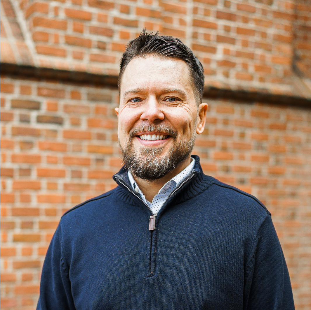 A man with short brown hair and a beard, wearing a navy sweater over a collared shirt, smiles while standing in front of a brick wall at Brightergy Renewable Energy.