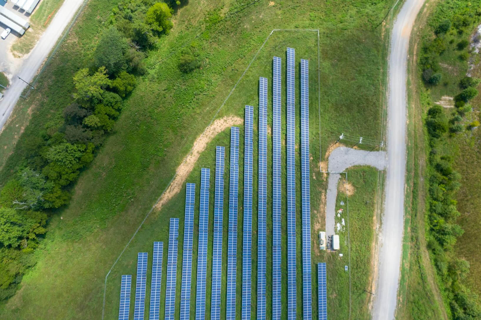 Aerial view of an array of solar panels, funded by nonprofit solar grants, arranged in parallel rows on a grassy field near trees and a paved road.