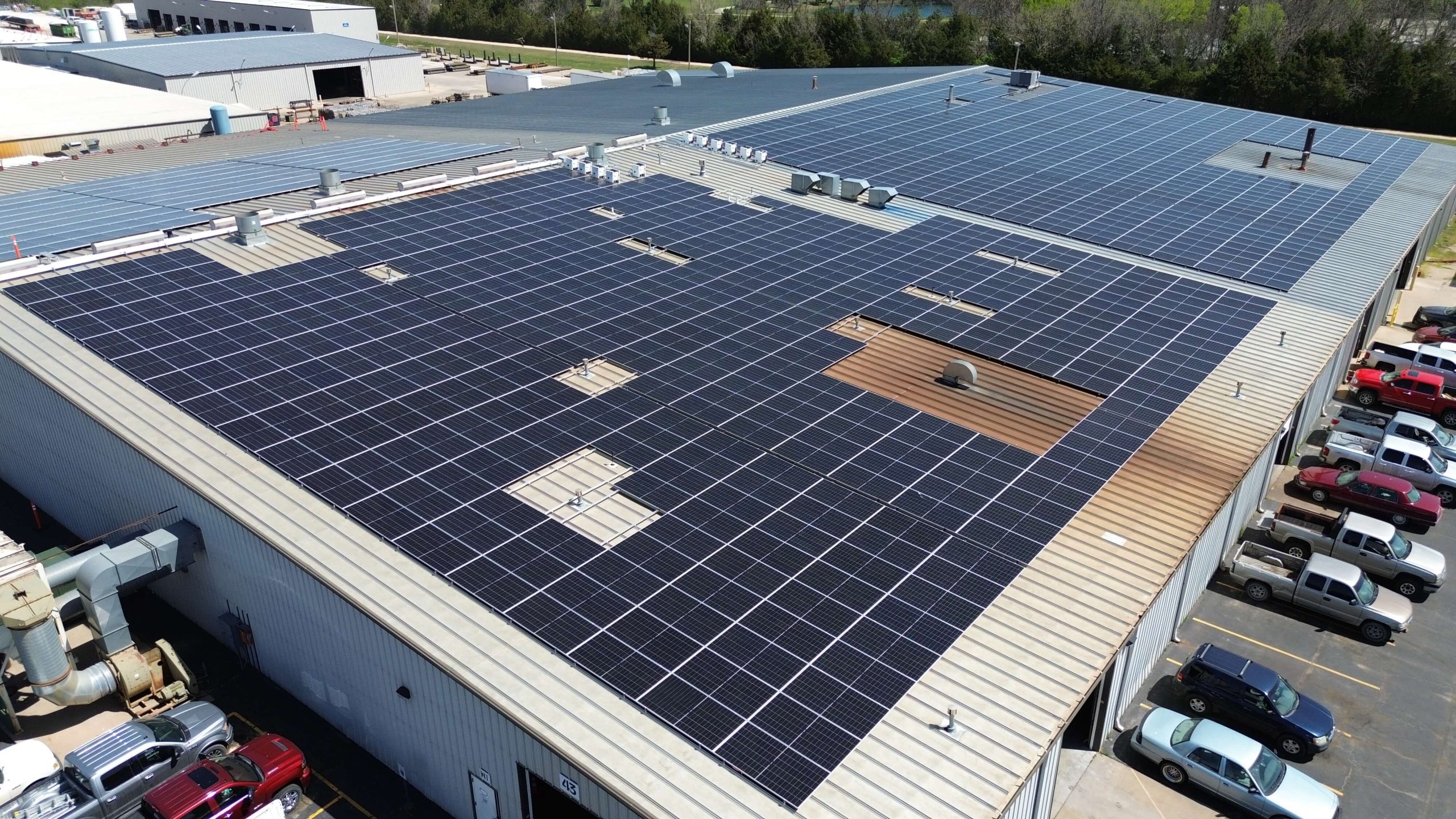 A large industrial building roof covered with solar panels, with several skylights and rooftop equipment visible, and cars parked along the side.